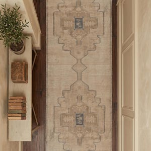 Puede incluir: Una alfombra pasillera larga y rectangular con una base de color beige y tostado descolorido, con un patrón geométrico de medallones repetidos delineados en azul apagado. La alfombra tiene un aspecto desgastado con flecos en los extremos.