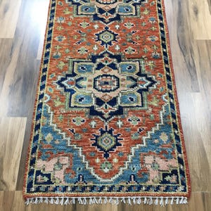 K&ouml;nnte beinhalten: Ein rechteckiger Teppich mit einem detaillierten Muster in Orange, Blau und Beige. Der Teppich hat ein sich wiederholendes geometrisches Design und einen blauen Rand. Wei&szlig;e Fransen s&auml;umen die Kanten. Er liegt auf einem Holzboden. Die Ma&szlig;e des Teppichs sind in Zentimetern.