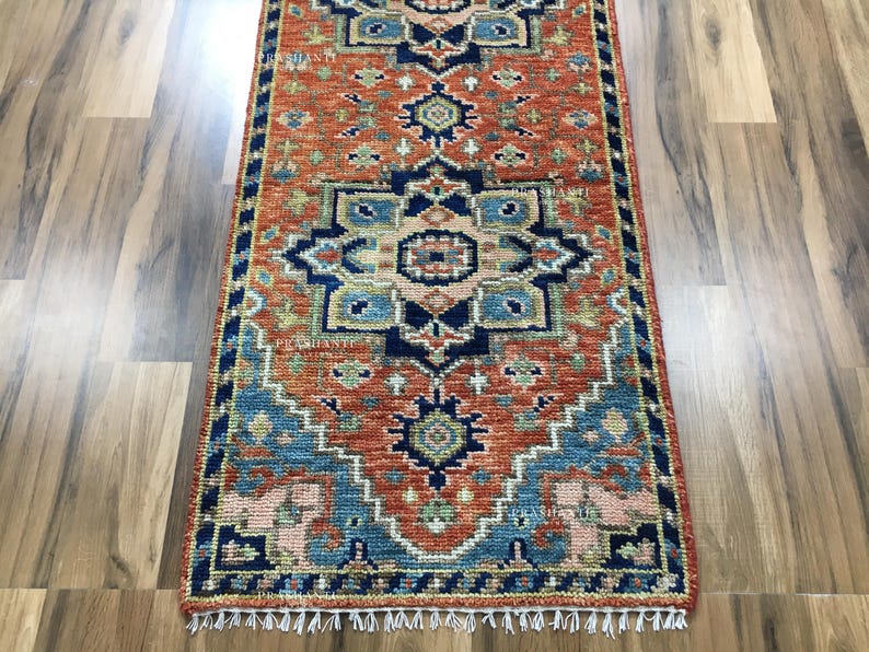 K&ouml;nnte beinhalten: Ein rechteckiger Teppich mit einem detaillierten Muster in Orange, Blau und Beige. Der Teppich hat ein sich wiederholendes geometrisches Design und einen blauen Rand. Wei&szlig;e Fransen s&auml;umen die Kanten. Er liegt auf einem Holzboden. Die Ma&szlig;e des Teppichs sind in Zentimetern.