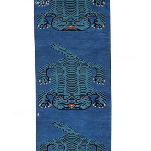 Könnte beinhalten: Ein rechteckiger blauer Teppich mit drei stilisierten Tigerdesigns. Jeder Tiger ist in Türkis und Schwarz dargestellt, mit einem roten Akzent im Gesicht. Der Teppich hat ein strukturiertes Aussehen, das auf eine gewebte Konstruktion hindeutet.