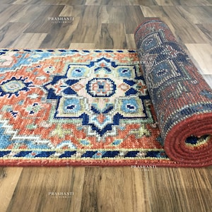 K&ouml;nnte beinhalten: Aufgerollter Teppich mit geometrischem Muster in Rot-, Blau- und Beiget&ouml;nen. Der Teppich ist teilweise ausgerollt und zeigt das komplizierte Design. Der Teppich liegt auf einem Holzboden.