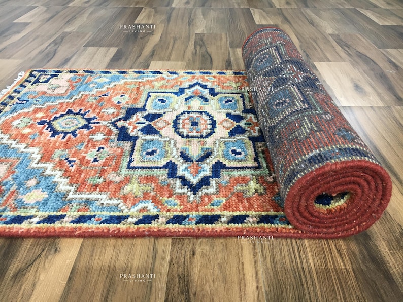 K&ouml;nnte beinhalten: Aufgerollter Teppich mit geometrischem Muster in Rot-, Blau- und Beiget&ouml;nen. Der Teppich ist teilweise ausgerollt und zeigt das komplizierte Design. Der Teppich liegt auf einem Holzboden.
