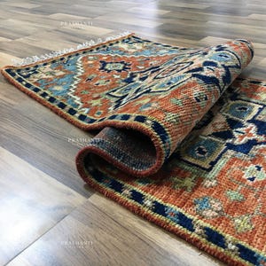 K&ouml;nnte beinhalten: Eine aufgerollte, rechteckige Teppich mit einem detaillierten geometrischen Muster in Orange-, Blau- und Marine-T&ouml;nen. Der Teppich hat einen Fransenrand und ist teilweise ausgerollt, wodurch das komplizierte Design sichtbar wird. Der Teppich liegt auf einem Holzboden.