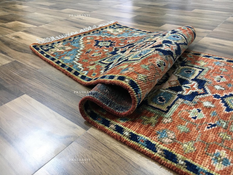 K&ouml;nnte beinhalten: Eine aufgerollte, rechteckige Teppich mit einem detaillierten geometrischen Muster in Orange-, Blau- und Marine-T&ouml;nen. Der Teppich hat einen Fransenrand und ist teilweise ausgerollt, wodurch das komplizierte Design sichtbar wird. Der Teppich liegt auf einem Holzboden.