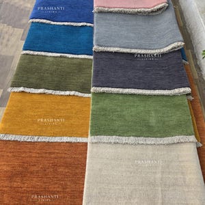 May include: A display of colorful rectangular rugs in various shades, including blue, pink, gray, green, and orange. Each rug has a fringed edge and the text "PRASHANTI LIVING" printed on it. The rugs are arranged side by side.