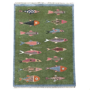 May include: A rectangular area rug with a deep olive green background featuring a repeating pattern of stylised fish in various colours including pink, blue, orange, and black. The rug has a blue patterned border and white fringe at both ends.