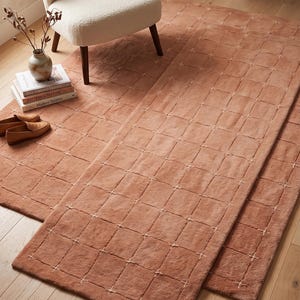 May include: A rectangular area rug in a solid terracotta colour featuring a grid pattern created by thin, light-coloured stitched lines. The rug has a soft, plush texture and is laid out flat on a light-coloured wooden floor surface.