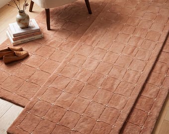 May include: A rectangular area rug in a solid terracotta color featuring a grid pattern created by thin, light-colored stitched lines. The rug has a soft, plush texture and is laid out flat on a light-colored wooden floor surface.