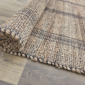 May include: A rolled-up jute rug, featuring a natural beige and black woven design. The rug's textured surface and rolled edge highlight the intricate weave. Partially unrolled, it displays its rectangular form and detailed pattern. The rug is suitable for any room.