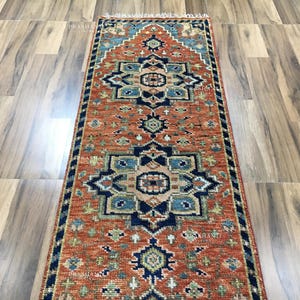 K&ouml;nnte beinhalten: Rechteckiger Teppich mit einem warmen, rot-orangen Hintergrund. Der Teppich zeigt aufwendige geometrische Muster in Marineblau, T&uuml;rkis und Gold. Der Teppich hat einen Fransenrand und eignet sich f&uuml;r Flure oder Eingangsbereiche.