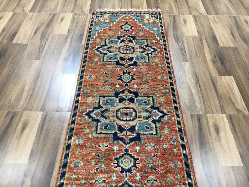 K&ouml;nnte beinhalten: Rechteckiger Teppich mit einem warmen, rot-orangen Hintergrund. Der Teppich zeigt aufwendige geometrische Muster in Marineblau, T&uuml;rkis und Gold. Der Teppich hat einen Fransenrand und eignet sich f&uuml;r Flure oder Eingangsbereiche.