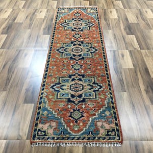 K&ouml;nnte beinhalten: Ein rechteckiger L&auml;uferteppich mit einem detaillierten Muster in Rost, Blau und Beige. Der Teppich hat ein sich wiederholendes geometrisches Design und einen gefransten Rand. Er wird auf einem Holzboden pr&auml;sentiert.