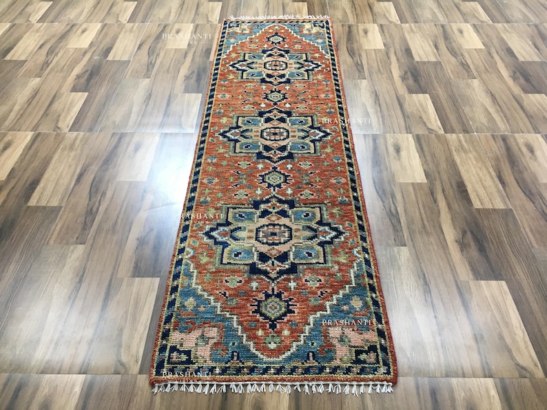 K&ouml;nnte beinhalten: Ein rechteckiger L&auml;uferteppich mit einem detaillierten Muster in Rost, Blau und Beige. Der Teppich hat ein sich wiederholendes geometrisches Design und einen gefransten Rand. Er wird auf einem Holzboden pr&auml;sentiert.