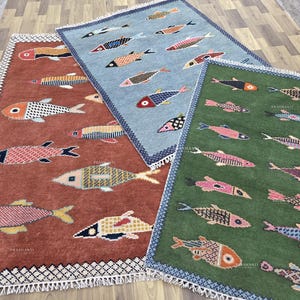 Pode incluir: Três tapetes retangulares com desenhos de peixes. Um é cor de ferrugem, outro azul claro e o terceiro verde. Cada tapete apresenta um padrão repetitivo de peixes coloridos, com franjas brancas.