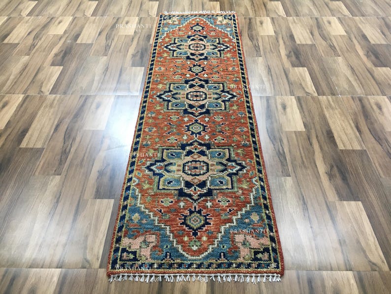 K&ouml;nnte beinhalten: Ein rechteckiger L&auml;uferteppich mit einem detaillierten Muster in Rost, Blau und Beige. Der Teppich hat ein sich wiederholendes geometrisches Design und einen gefransten Rand. Er wird auf einem Holzboden pr&auml;sentiert.
