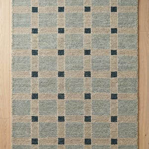 May include: A rectangular area rug featuring a grid pattern of light blue and beige squares with dark navy blue accent squares at each intersection. The rug has a textured surface and is finished with short, light beige fringe along the top and bottom edges.