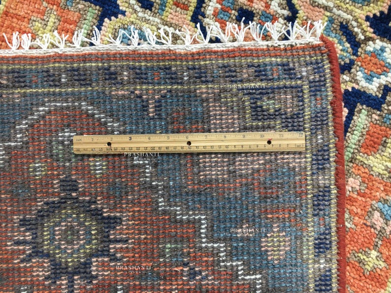 K&ouml;nnte beinhalten: Nahaufnahme eines Teils eines gemusterten Teppichs mit einem Lineal. Der Teppich weist eine Mischung aus Farben auf, darunter Blau, Orange und Rot, mit einem detaillierten geometrischen Design. Das Lineal misst in Zentimetern.