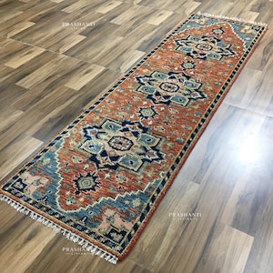 K&ouml;nnte beinhalten: Ein rechteckiger L&auml;uferteppich mit einem geometrischen Muster in Orange-, Blau- und Cremet&ouml;nen. Der Teppich hat einen Fransenrand und wird auf einem Holzboden pr&auml;sentiert. Das Design zeigt sich wiederholende Blumen- und geometrische Motive.