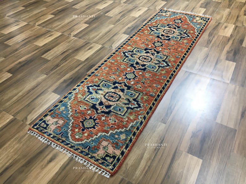 K&ouml;nnte beinhalten: Ein rechteckiger L&auml;uferteppich mit einem geometrischen Muster in Orange-, Blau- und Cremet&ouml;nen. Der Teppich hat einen Fransenrand und wird auf einem Holzboden pr&auml;sentiert. Das Design zeigt sich wiederholende Blumen- und geometrische Motive.