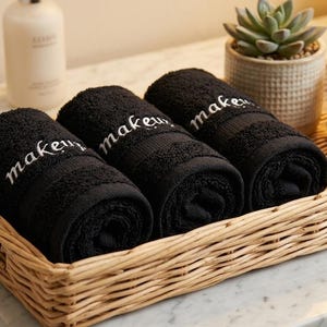Puede incluir: Tres toallas negras enrolladas con la palabra makeup bordada en letra blanca, dispuestas en una bandeja rectangular de mimbre trenzado junto a una pila de tres toallas blancas dobladas y una pequeña planta suculenta en una maceta de cerámica.
