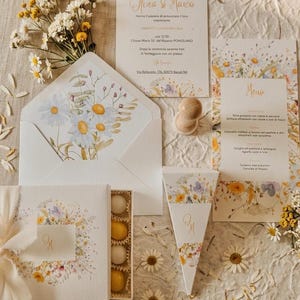 Könnte beinhalten: Ein Hochzeitspapeterie-Set mit weißer Einladung, Menükarte und Umschlag, verziert mit Aquarell-Illustrationen von Gänseblümchen und Wildblumen. Enthält eine Schachtel mit gelben und weißen Bonbons, eine dreieckige Schachtel und einen Holzstempel.