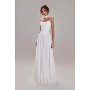 May include: A floor-length white gown featuring a strapless bodice with a draped cowl neckline, a fitted corset-style waist, and a flowing skirt. The garment includes a matching white choker accessory worn around the neck.