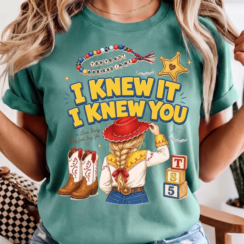 May include: A teal t-shirt featuring a graphic print with the text "I KNEW IT I KNEW YOU" in yellow. The design includes a beaded necklace, a yellow sheriff star, a blonde character in a red cowboy hat, brown boots, and three alphabet blocks.