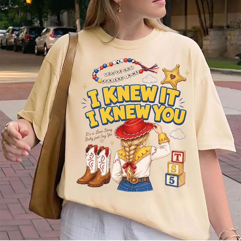 May include: A cream-colored t-shirt featuring a graphic print with the text "I KNEW IT I KNEW YOU," a beaded bracelet with the text "YOU'VE GOT A FRIEND IN ME," a cowboy hat, boots, a person with a braided ponytail, and blocks labeled "T S 5."