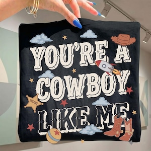 May include: A black t-shirt featuring the white text "YOU'RE A COWBOY LIKE ME" surrounded by illustrations of a brown cowboy hat, a rocket ship, a gold sheriff star badge, blue clouds, small stars, a striped ball, and brown cowboy boots.