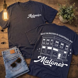 May include: A dark navy blue short-sleeve t-shirt featuring a white script logo on the front chest. The back displays a white line-art illustration of a building storefront with the text "BEST BURGERS IN HASTINGS, MA!" and "Malone's" below.