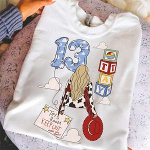 K&ouml;nnte beinhalten: Ein wei&szlig;es Sweatshirt mit Rundhalsausschnitt und einer Grafik einer Person mit langem blonden Haar, die eine Jacke mit Kuhfleckenmuster und roten Fransen tr&auml;gt. Das Design umfasst einen blauen, wolkenmusterartigen Luftballon in Form der 13, Alphabet-Bl&ouml;cke mit der Aufschrift TAY und ein Schild mit dem Text TAYS ROOM KEEP OUT except you Travis.