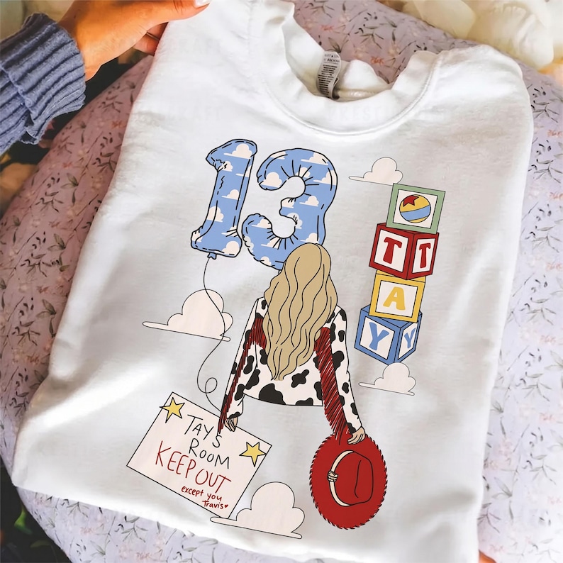 K&ouml;nnte beinhalten: Ein wei&szlig;es Sweatshirt mit Rundhalsausschnitt und einer Grafik einer Person mit langem blonden Haar, die eine Jacke mit Kuhfleckenmuster und roten Fransen tr&auml;gt. Das Design umfasst einen blauen, wolkenmusterartigen Luftballon in Form der 13, Alphabet-Bl&ouml;cke mit der Aufschrift TAY und ein Schild mit dem Text TAYS ROOM KEEP OUT except you Travis.