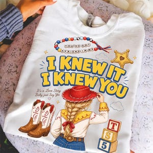 May include: A white crewneck sweatshirt featuring a graphic print of a person with blonde braided hair wearing a red cowboy hat. The text reads I KNEW IT I KNEW YOU, YOU'VE GOT A FRIEND IN ME, and It's a Love Story Baby just Say Yes.