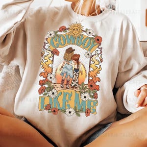 May include: A cream-coloured crewneck sweatshirt featuring a graphic print of two figures in cowboy hats and boots standing together. The design includes the text "COWBOY LIKE ME" in blue, surrounded by orange flames, flowers, and a sun icon.