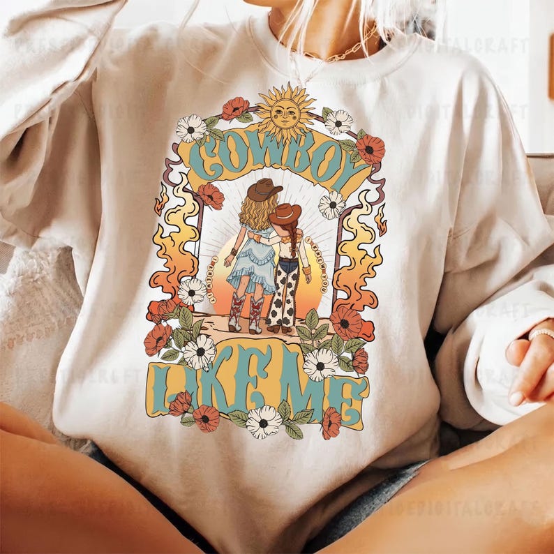 May include: A cream-coloured crewneck sweatshirt featuring a graphic print of two figures in cowboy hats and boots standing together. The design includes the text "COWBOY LIKE ME" in blue, surrounded by orange flames, flowers, and a sun icon.