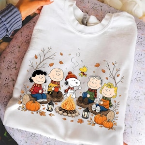 May include: A white crewneck sweatshirt featuring a graphic illustration of the Peanuts characters sitting around a campfire. The design includes Snoopy, Charlie Brown, Lucy, Linus, and Sally wearing scarves, surrounded by autumn leaves and pumpkins.