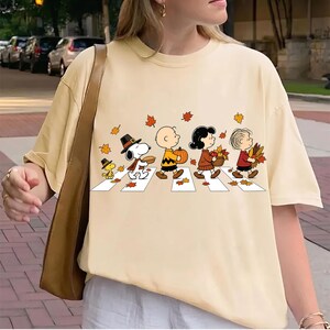 Puede incluir: Una camiseta de manga corta color crema con un gráfico de los personajes de Peanuts caminando por un paso de cebra. El diseño incluye a Snoopy, Woodstock, Charlie Brown, Lucy y Linus cargando elementos otoñales como calabazas y hojas.