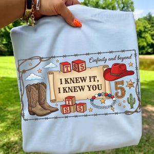 Pu&ograve; includere: Una t-shirt azzurro chiaro con una stampa grafica raffigurante una pergamena con il testo "I KNEW IT, I KNEW YOU", blocchi "TS5", un cappello da cowboy rosso, stivali marroni, un braccialetto di perline, un cactus, una stella e "Cinfinity and beyond".