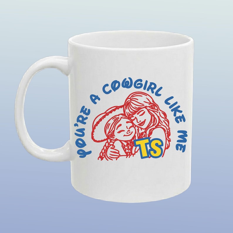 Op de afbeelding: Een witte keramische mok met een rode lijntekening van twee mensen die elkaar omhelzen, met de blauwe tekst "YOU'RE A COWGIRL LIKE ME" in een boog erboven en de gele letters "TS" prominent geplaatst bij de onderkant van het ontwerp.