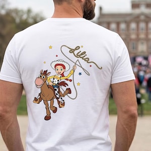 May include: A white t-shirt featuring a graphic of a cowgirl character riding a brown horse. The character wears a white hat, yellow shirt, and cow-print vest. A lasso is held above, and the name Lilia is written in cursive script with stars.