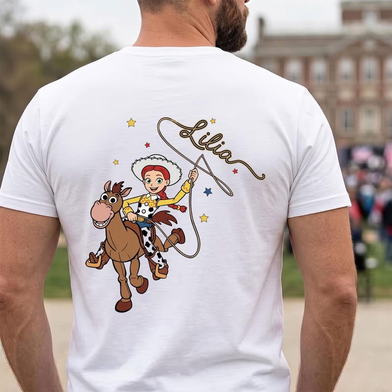 May include: A white t-shirt featuring a graphic of a cowgirl character riding a brown horse. The character wears a white hat, yellow shirt, and cow-print vest. A lasso is held above, and the name Lilia is written in cursive script with stars.
