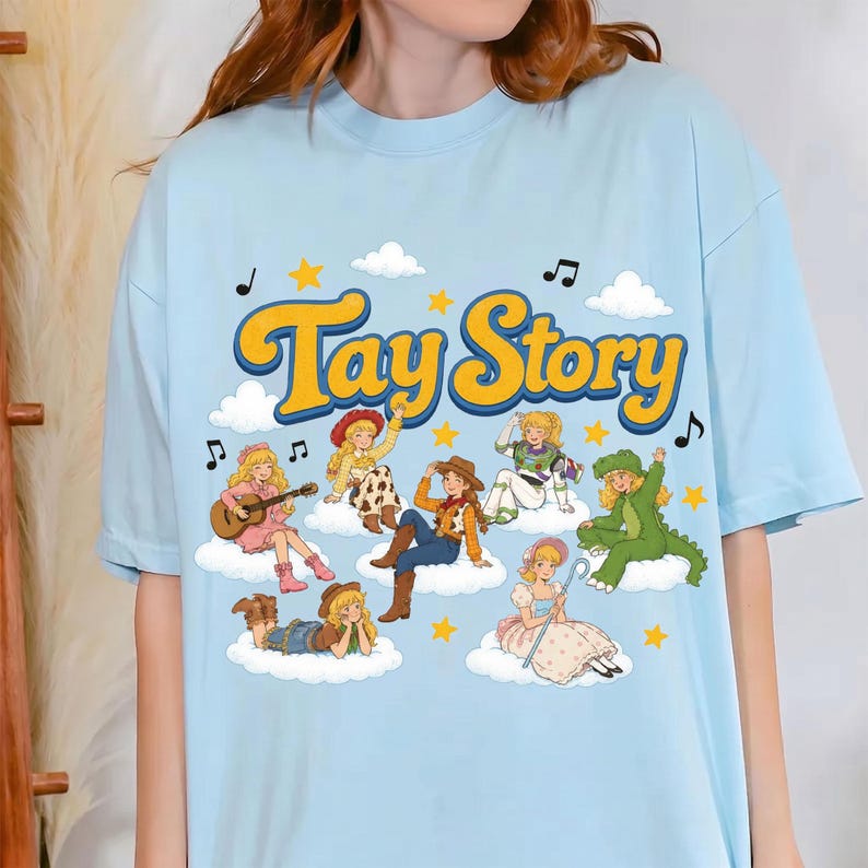 May include: A light blue t-shirt featuring the yellow text "Tay Story" above a graphic of multiple blonde characters dressed in various costumes, including a cowgirl, a space ranger, and a green dinosaur, all resting on white clouds with stars.