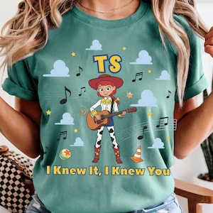 Peut inclure: Un t-shirt bleu sarcelle avec une illustration d'une cow-girl jouant de la guitare acoustique. Le motif comprend des nuages bleus, des &eacute;toiles jaunes, des notes de musique, une balle, un c&ocirc;ne de signalisation et le texte TS et I Knew It, I Knew You.