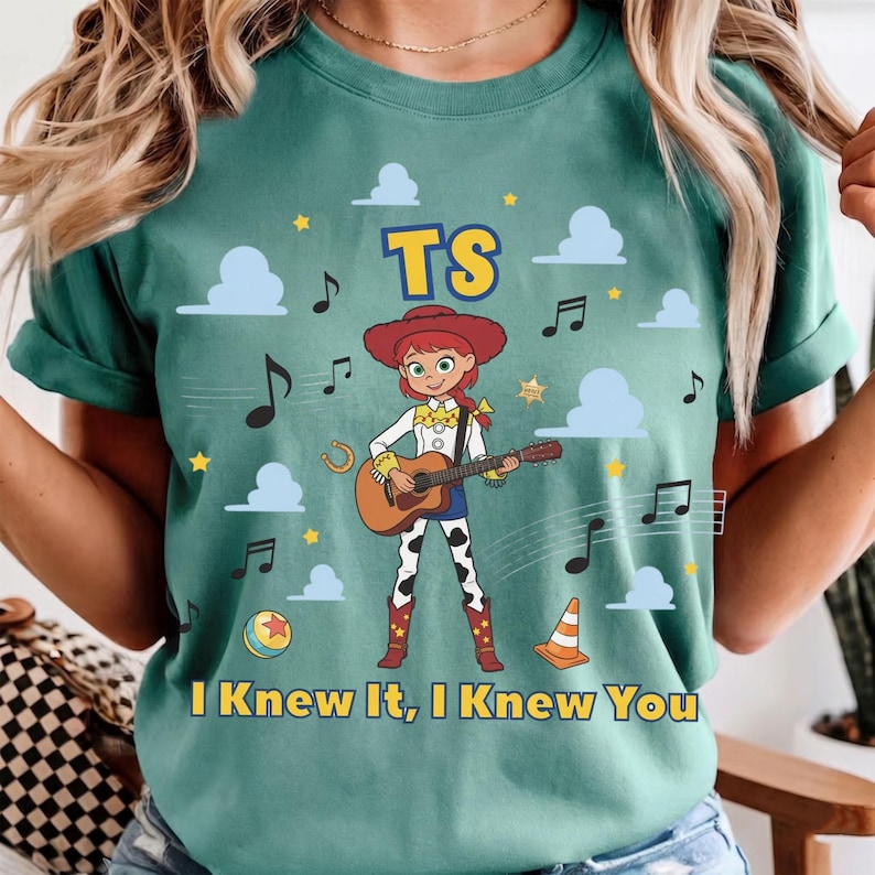 Peut inclure: Un t-shirt bleu sarcelle avec une illustration d'une cow-girl jouant de la guitare acoustique. Le motif comprend des nuages bleus, des &eacute;toiles jaunes, des notes de musique, une balle, un c&ocirc;ne de signalisation et le texte TS et I Knew It, I Knew You.