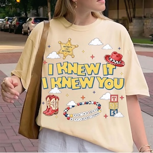 Op de afbeelding: Een crèmekleurig T-shirt met korte mouwen en een grafische print met de tekst I KNEW IT I KNEW YOU. Het ontwerp bevat een gele sheriffster, een rode cowboyhoed, rode cowboylaarzen, een kralenarmband met de tekst You've got a friend in me, en gestapelde alfabetblokken met de letters T, S en 5, omringd door wolken en sterren.
