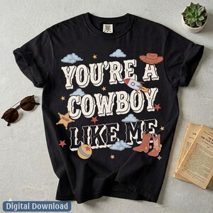May include: A black short-sleeve t-shirt featuring the white text "YOU'RE A COWBOY LIKE ME" surrounded by graphic illustrations of a brown cowboy hat, a rocket ship, a sheriff star badge, a colorful ball, and brown cowboy boots with stars.