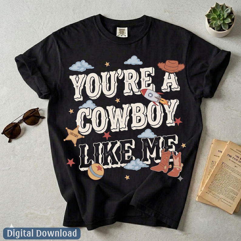 May include: A black short-sleeve t-shirt featuring the white text "YOU'RE A COWBOY LIKE ME" surrounded by graphic illustrations of a brown cowboy hat, a rocket ship, a sheriff star badge, a colorful ball, and brown cowboy boots with stars.