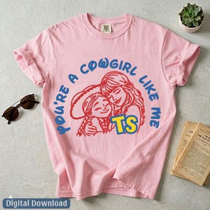 Op de afbeelding: Een lichtroze T-shirt met korte mouwen voorzien van een afbeelding van twee figuren in een rode omtrek, met de blauwe tekst "YOU'RE A COWGIRL LIKE ME" in een boog erboven en de gele tekst "TS" eronder op de voorzijde.