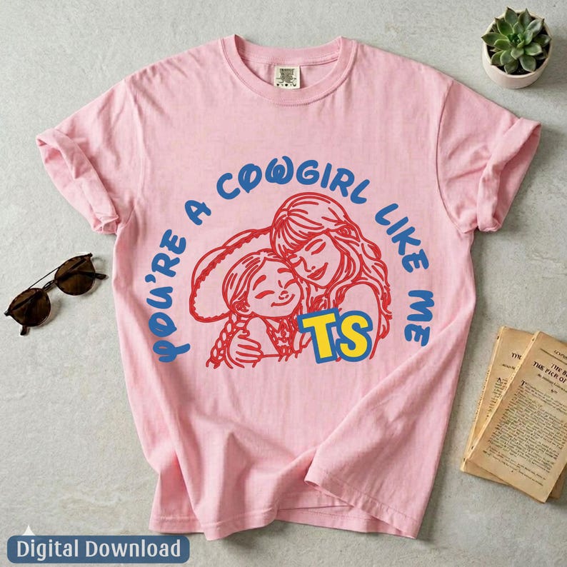 Op de afbeelding: Een lichtroze T-shirt met korte mouwen voorzien van een afbeelding van twee figuren in een rode omtrek, met de blauwe tekst "YOU'RE A COWGIRL LIKE ME" in een boog erboven en de gele tekst "TS" eronder op de voorzijde.