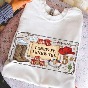 Pu&ograve; includere: Una t-shirt bianca con una stampa grafica di una pergamena con il testo "I KNEW IT, I KNEW YOU", circondata da stivali da cowboy, blocchi rossi "TS 5", un cappello da cowboy rosso, un numero 5, un cactus e un braccialetto di perline.